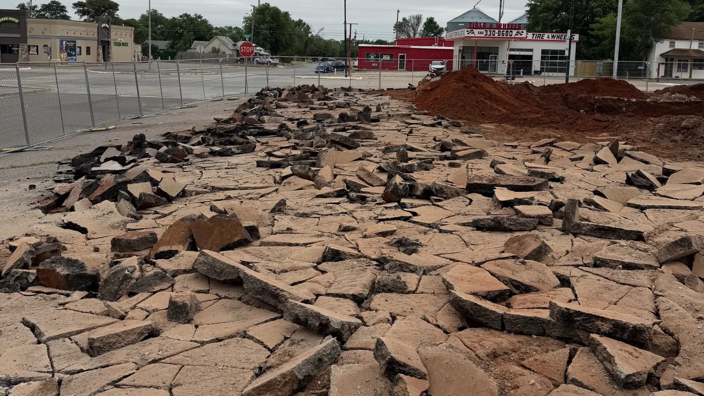 A commercial parking lot broken into loose slabs of asphalt and concrete across the exposed sand subgrade, fenced off from the street