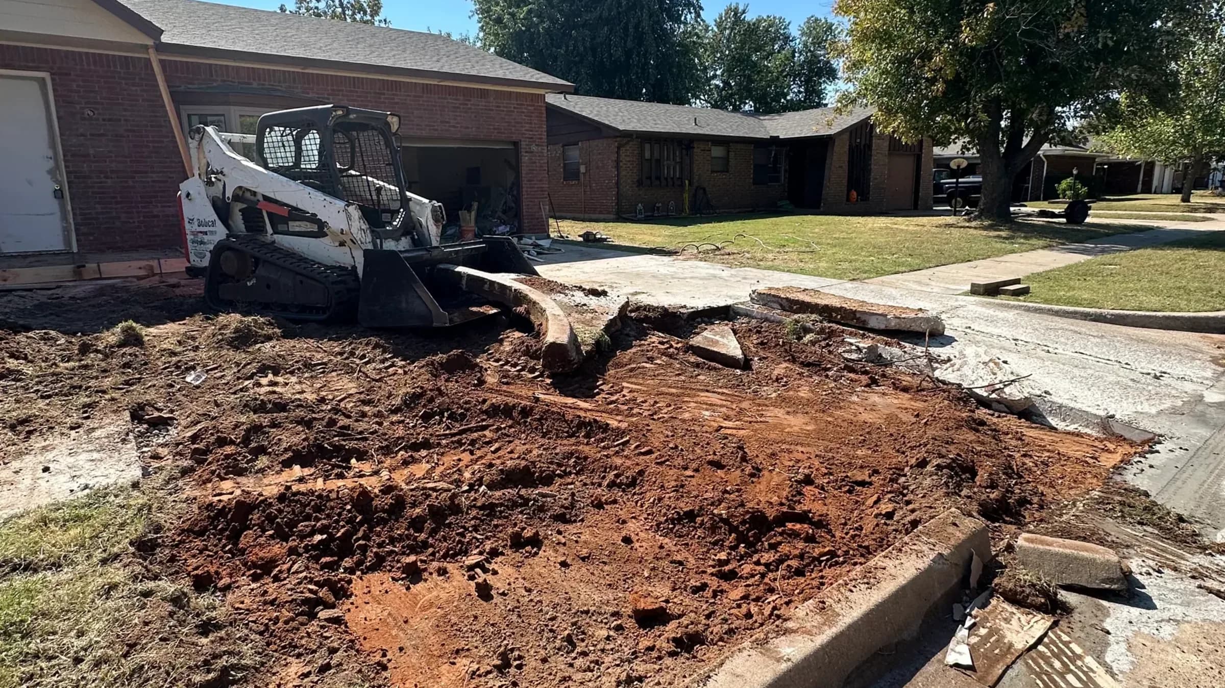 A compact track loader breaking a concrete driveway out at the curb line, broken slab sections and red dirt where the drive used to run