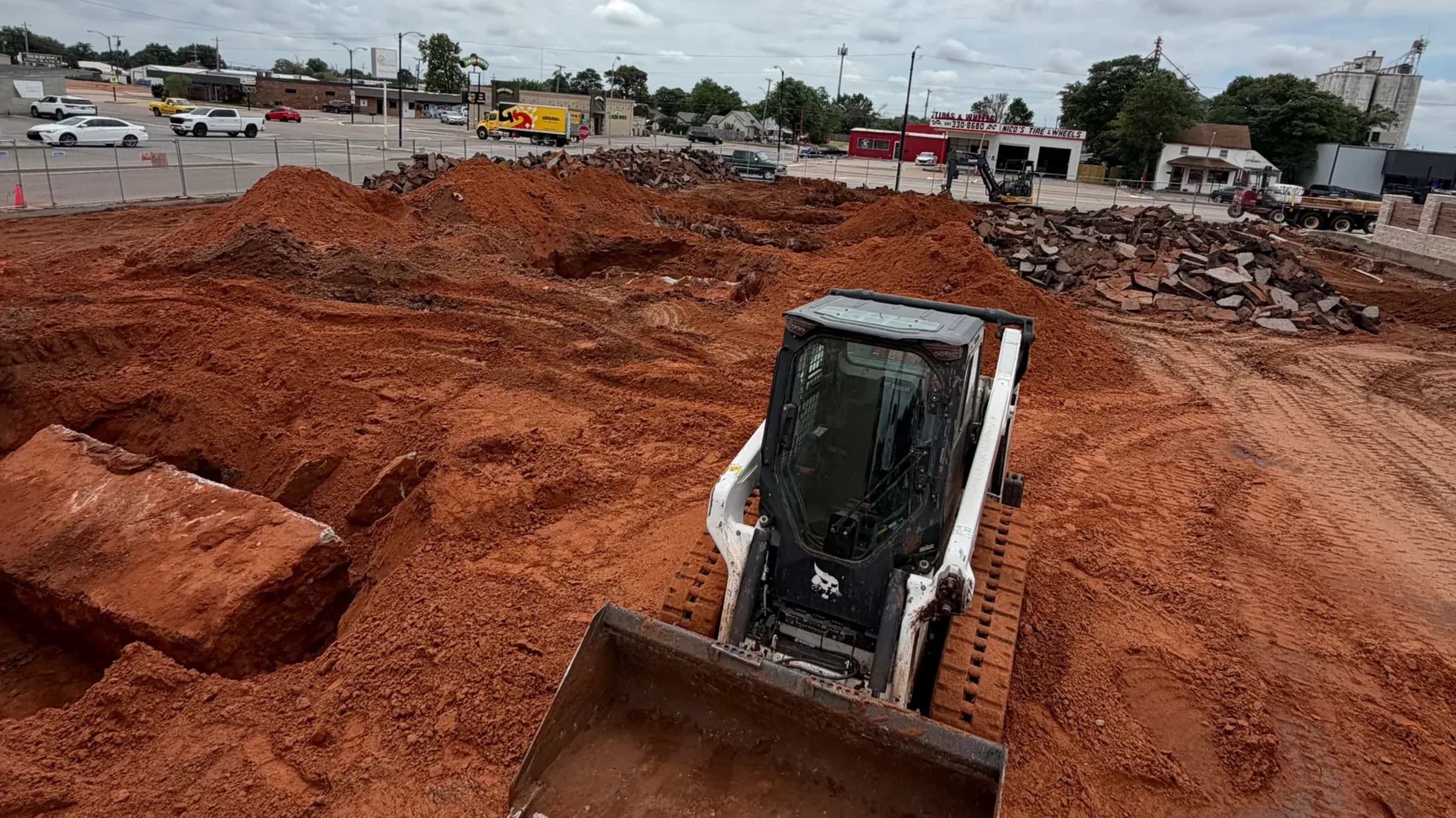A compact track loader with a toothed bucket working a cleared commercial lot, spoil piles of red clay and broken concrete stacked along the back fence