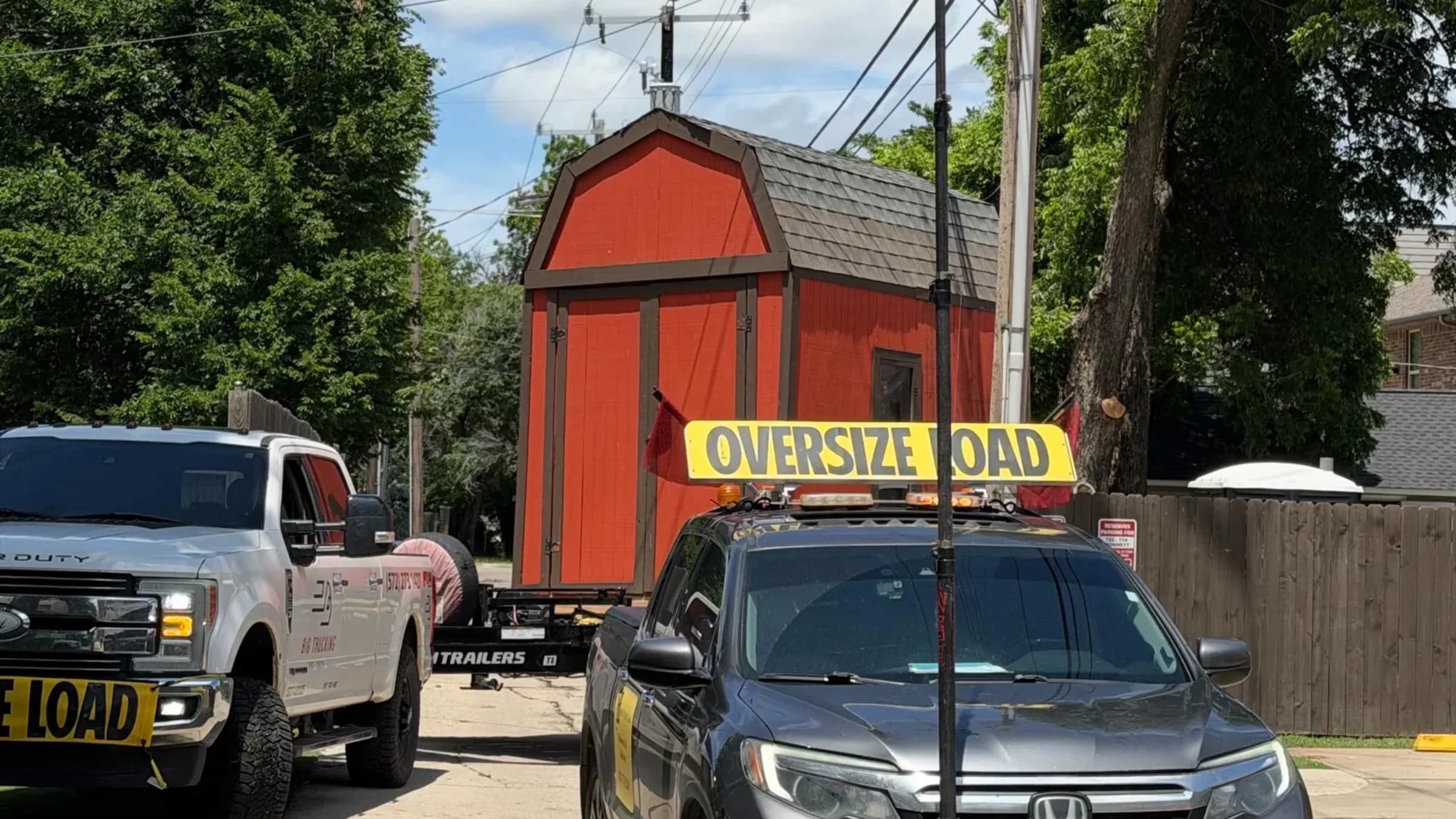 A storage shed strapped to a gooseneck trailer moving down a residential street behind an escort vehicle carrying an oversize load banner