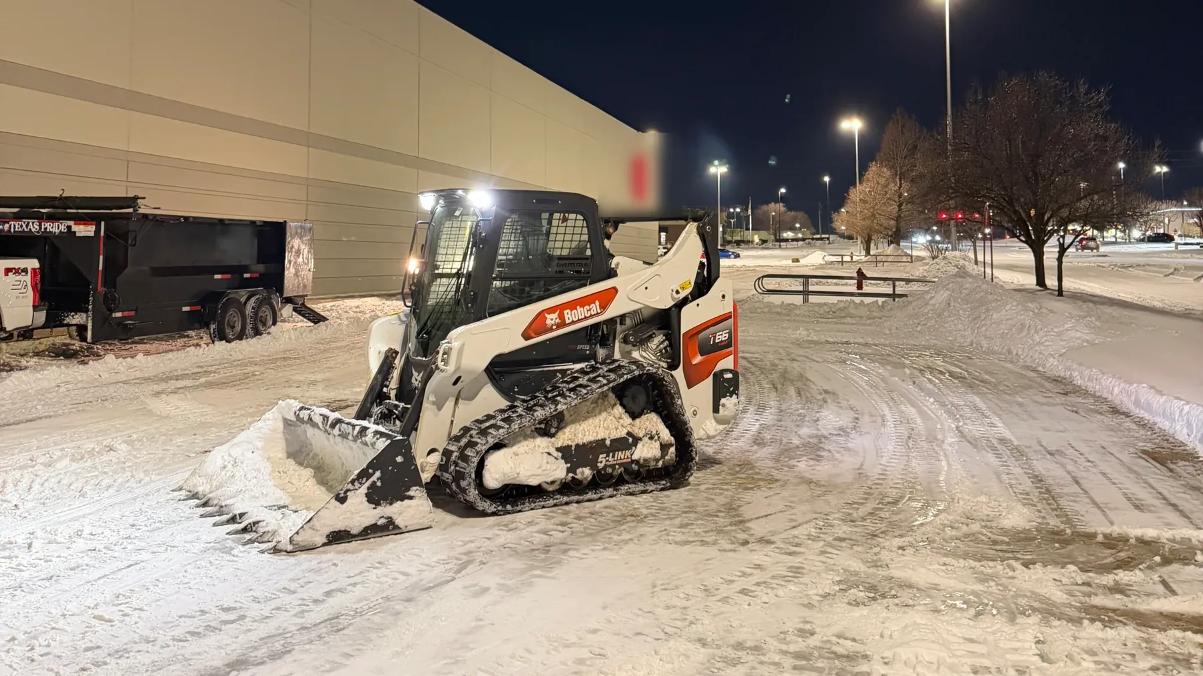 A compact track loader pushing snow across a lit commercial parking lot after dark, with cleared lanes and snow banked along the edges